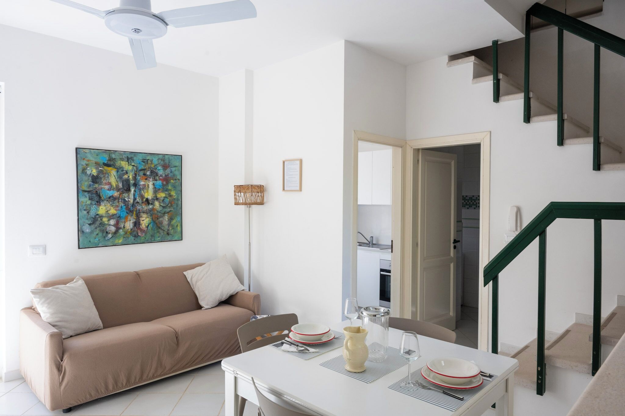 Una sala de estar moderna con un sofá, una mesa de comedor y obras de arte. Se ve una escalera con barandales verdes.