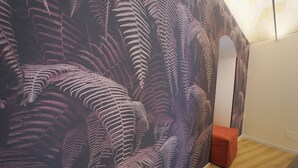 Ein Zimmer mit einer großen Wandmalerei von Farnblättern, Holzboden und einem kleinen, orangefarbenen Hocker.