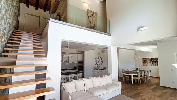 A spacious living area with a modern staircase, a dining area with a wooden table and chairs, and a kitchen in the background.