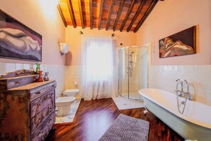A bathroom with a wooden floor, a white bathtub, a glass shower enclosure, a white toilet, and a vintage dresser. - Holiday Home Croce (Lucignano)