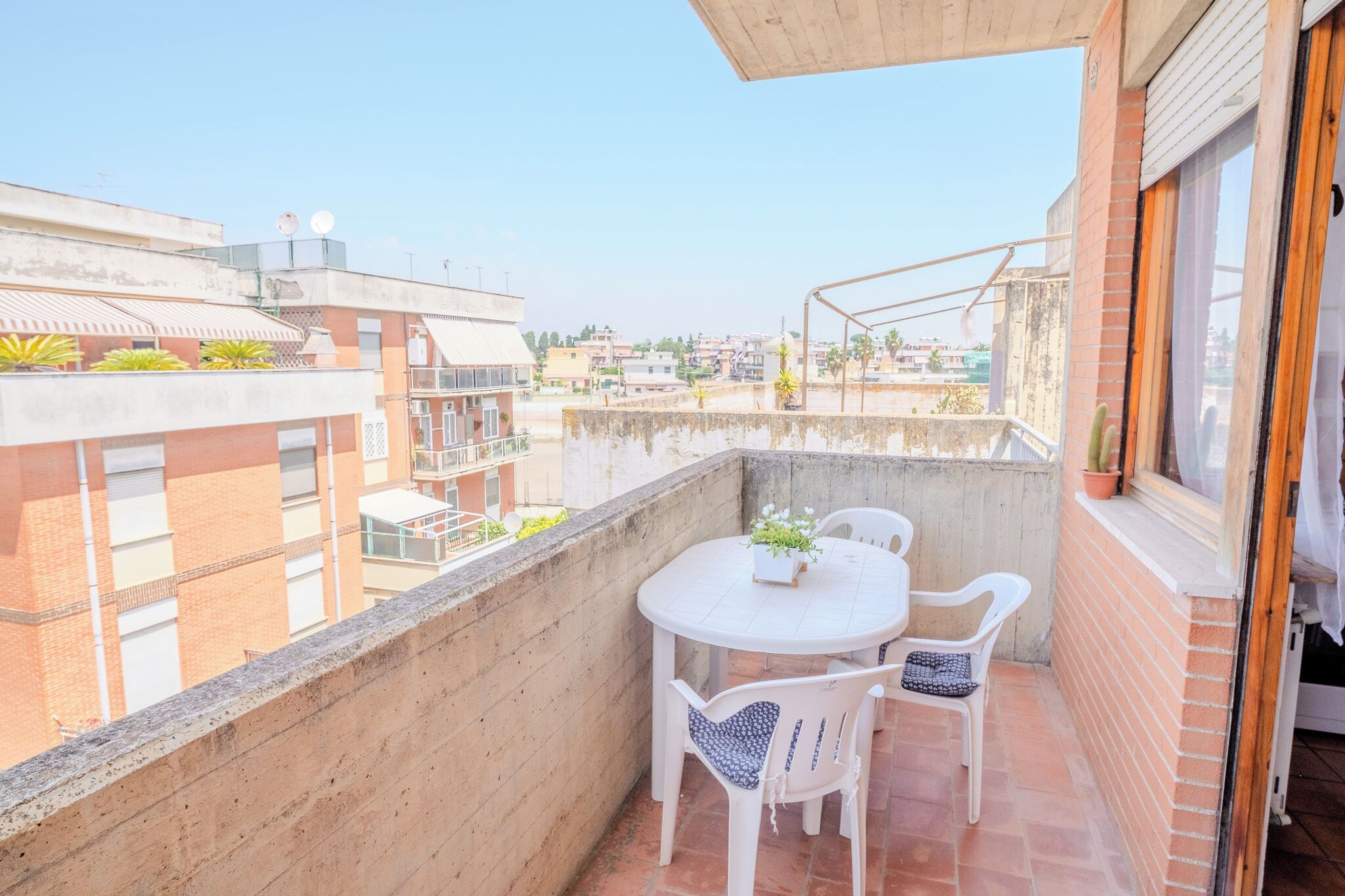 A balcony with a round table and two chairs, a potted plant, and a view of neighboring buildings.