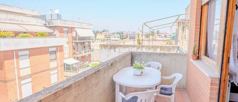 A balcony with a round table and two chairs, a potted plant, and a view of neighboring buildings.