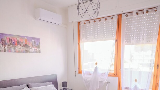 A bedroom with a bed, a nightstand, an air conditioner, a window with curtains, and a radiator. - San Giacomo Sweet Home (Nettuno)
