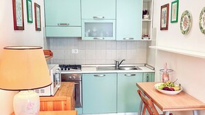 A compact kitchen with teal cabinets, a gas stove, and a sink. There's a dining table with a bowl of fruit and a lamp on a wooden side table. - La Casina Di Giuliana (Principina a Mare)