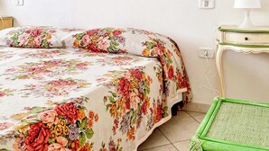 A room with a floral bedspread, a painting of cherubs, a mirror, a lamp, and a green chair. - La Casina Di Giuliana (Principina a Mare)