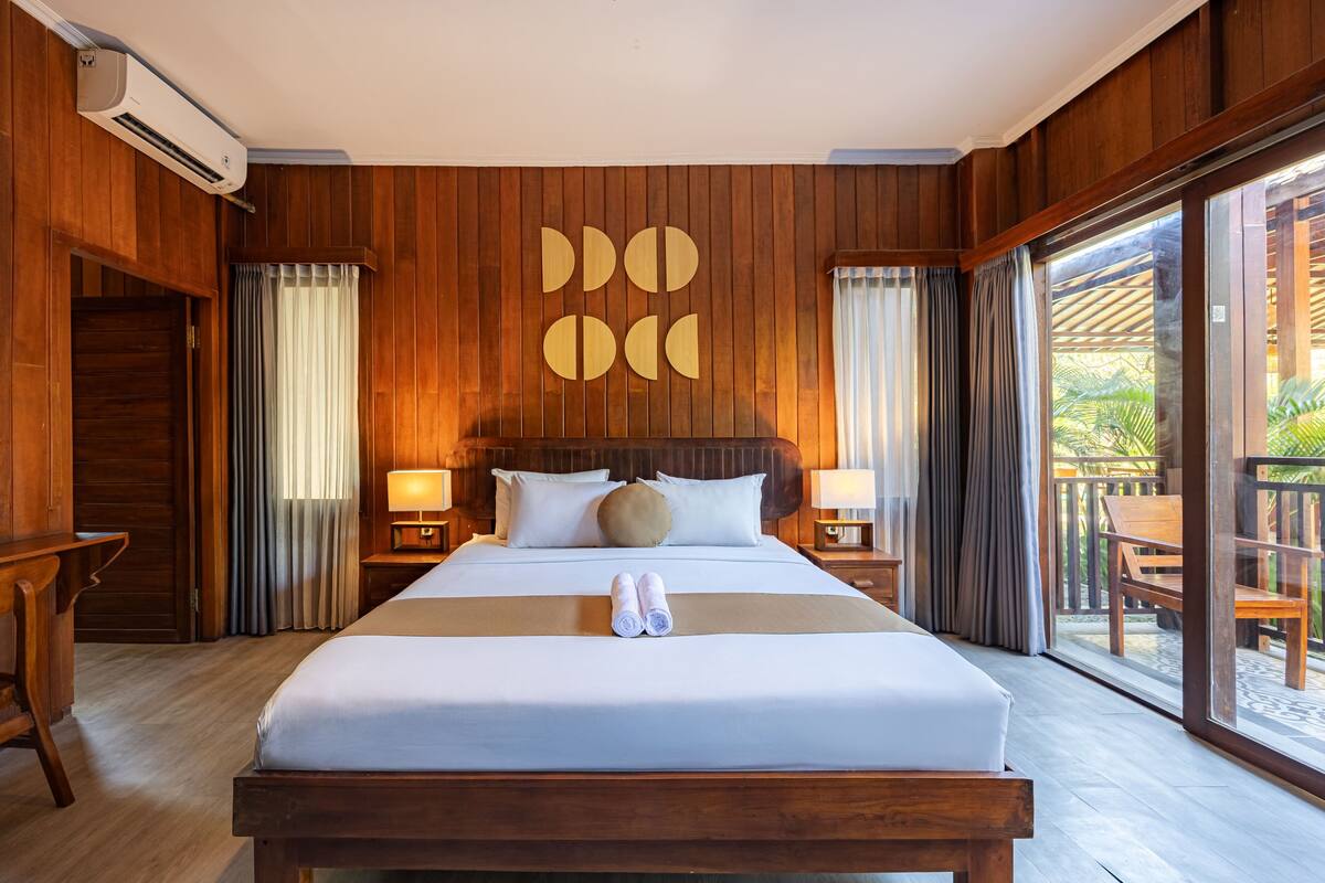 A hotel room with a wooden paneled wall, a large bed with white linens, two bedside lamps, and a view through a glass door to the outside.