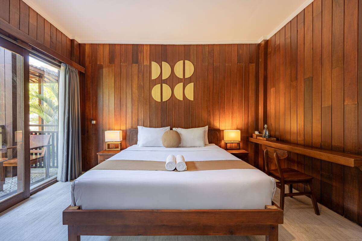 A large bed with a wooden headboard, two bedside tables with lamps, a bench, and a view of the outdoors through a sliding door.