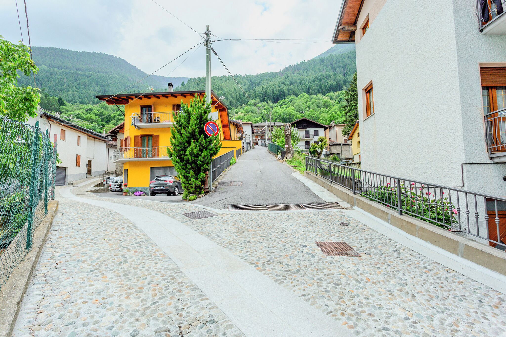 A cobblestone street in a village with houses on either side, a yellow building with a balcony, and a mountain in the background.