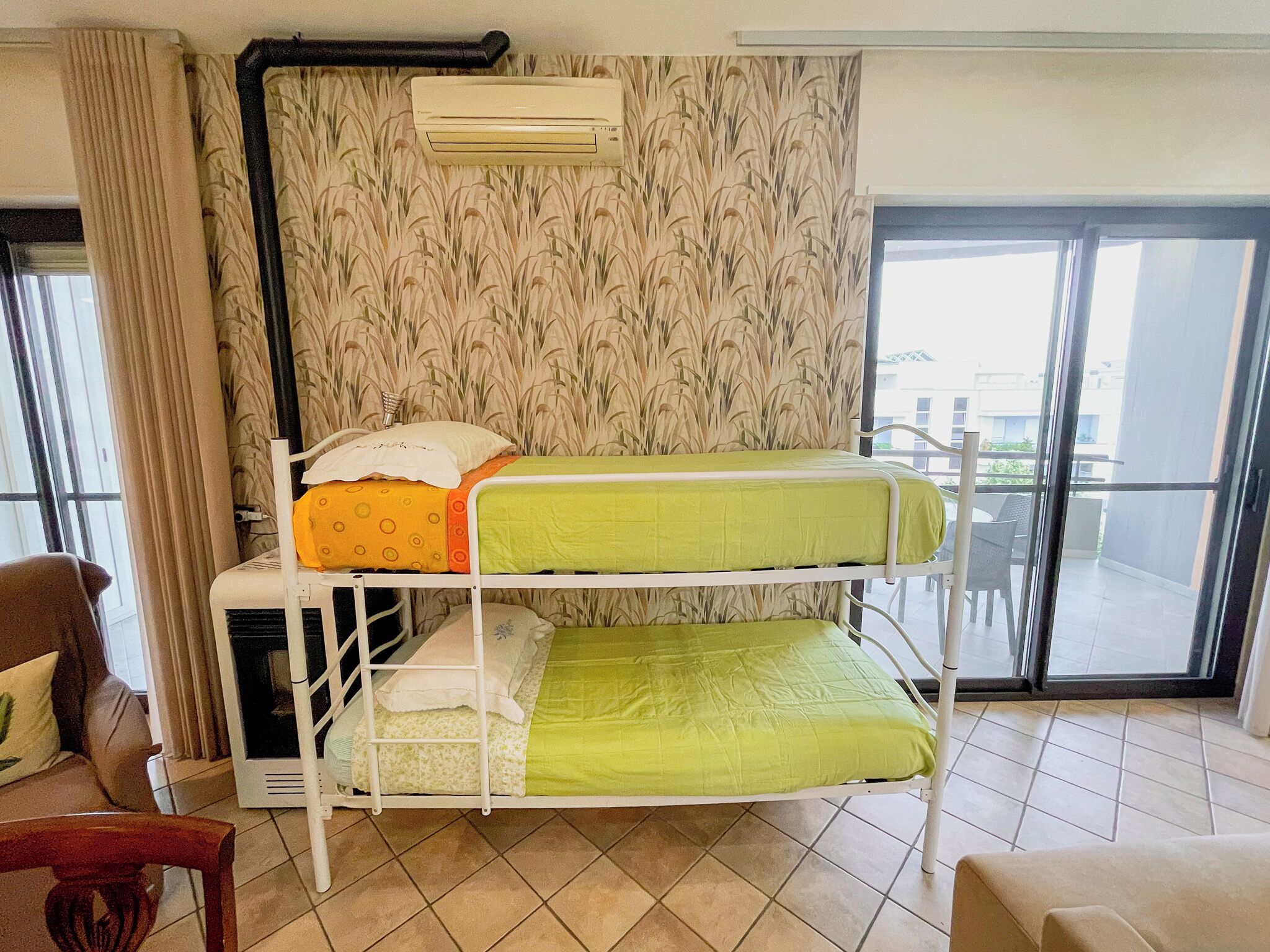Une chambre avec un lit superposé, une fenêtre avec des rideaux, un climatiseur fixé au mur et une vue sur le balcon extérieur.