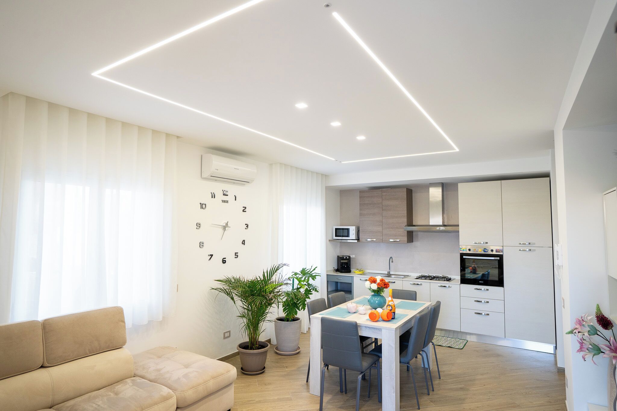 A modern living room with a sofa, dining table, and chairs. There is a kitchen area with cabinets, a microwave, and a stove.