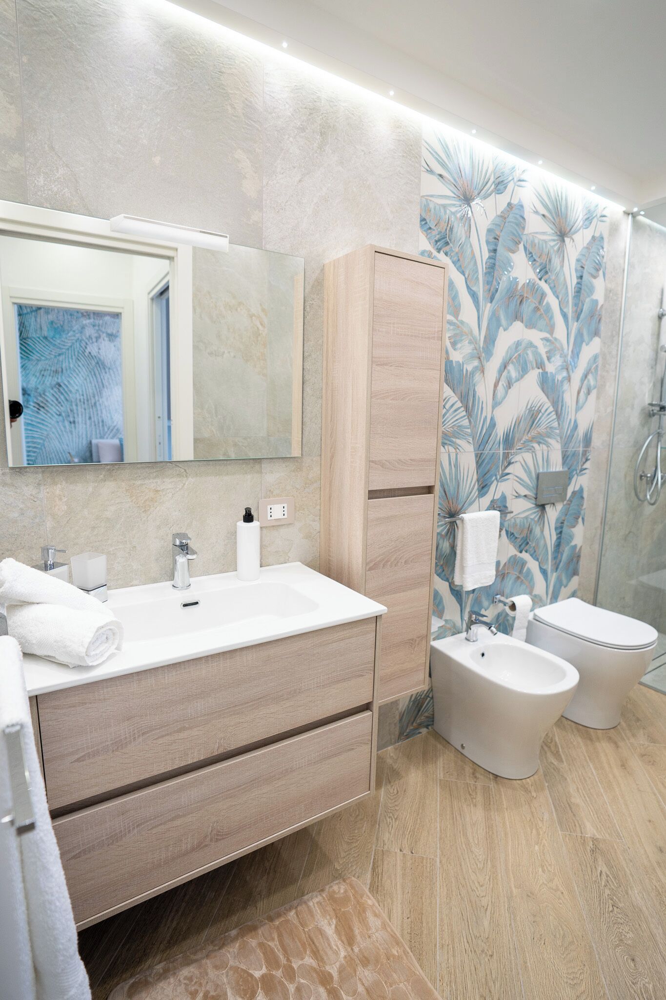 Une salle de bain moderne avec un grand miroir, un lavabo blanc, un meuble en bois et un espace douche agrémenté d’un papier peint aux motifs tropicaux.