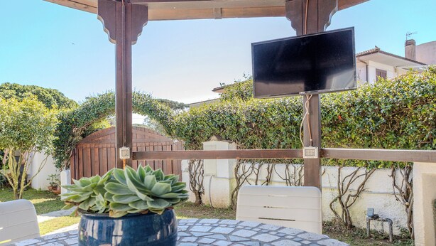A covered outdoor seating area with a potted succulent plant, a table, and a TV screen. - Villa Magnolia (Sabaudia)