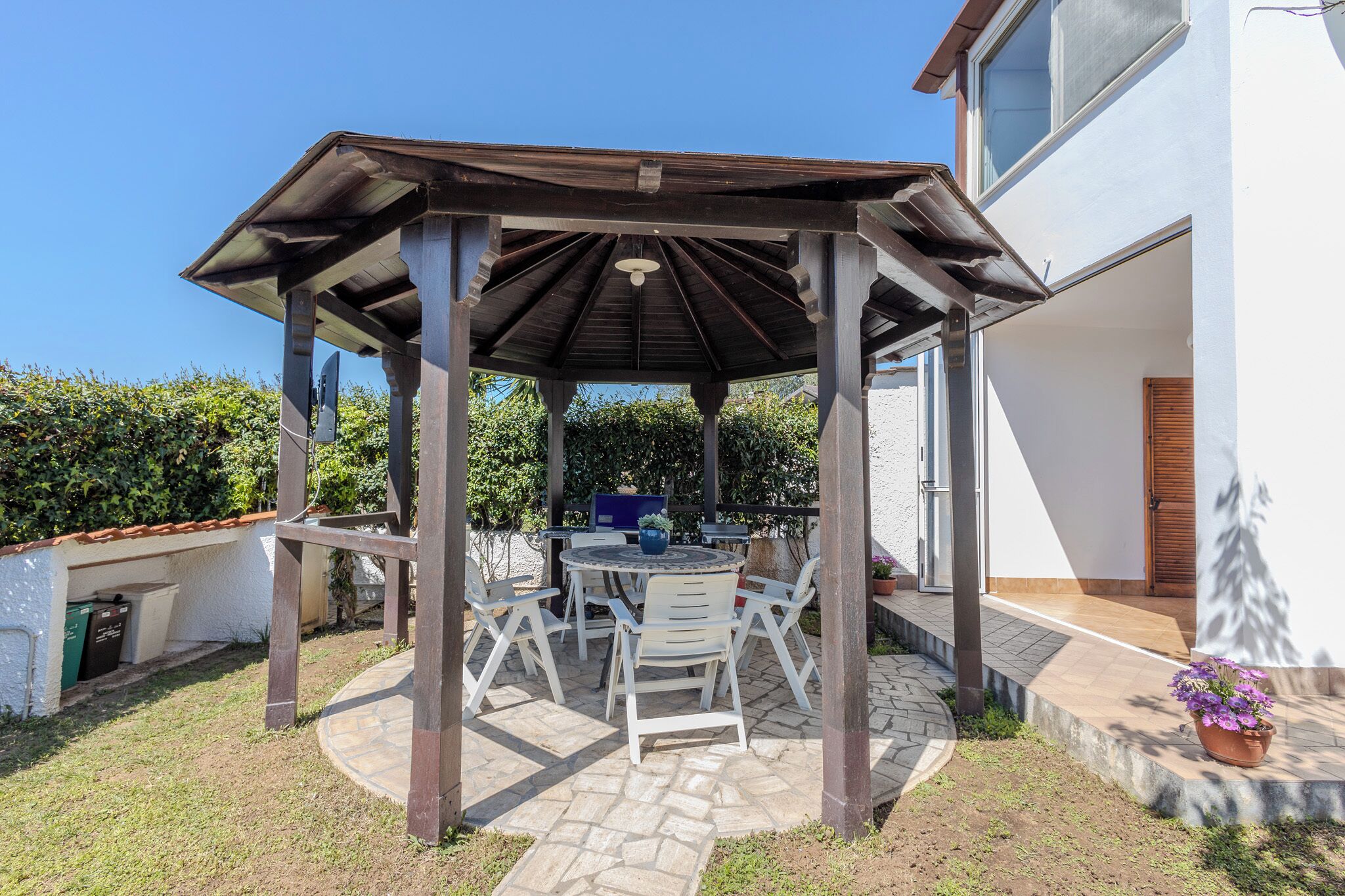 En täckt uteplats med ett bord och stolar, en pergola och utsikt över ett hus.