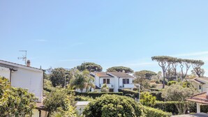 A white villa with a tiled roof, surrounded by lush greenery and trees, set in a residential area with other houses visible in the background. - Villa Magnolia (Sabaudia)