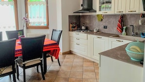 A kitchen with white cabinets, a gas stove, and a sink. There is a dining table with chairs and a window with heart-patterned curtains. - Casa TreSoli - Baselga di Pinè (Baselga di Piné)