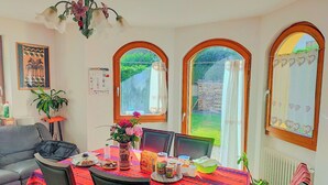 A dining area with a table set for two, a laptop, and various items on the table. There are two chairs, a sofa, and a potted plant. The room has arched windows with curtains, a chandelier, and a calendar on the wall. - Casa TreSoli - Baselga di Pinè (Baselga di Piné)