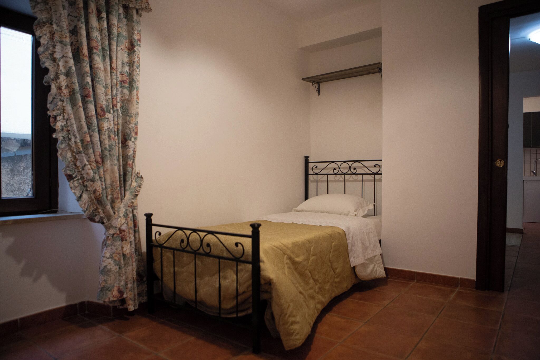 A small, single-bed room with a metal headboard, a window with floral curtains, and a tiled floor.