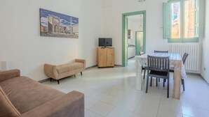 Una sala de estar con un sofá, un televisor sobre un mueble de madera, una mesa de comedor con sillas y una ventana con contraventanas verdes.