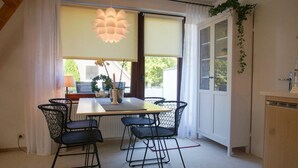 Un comedor con una mesa y sillas, una lámpara colgante, una ventana con cortinas y vistas al exterior con vegetación.