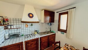 A compact kitchen with a gas stove, a microwave, wooden cabinets, and a window with curtains. - Casa Puni (Olbia)