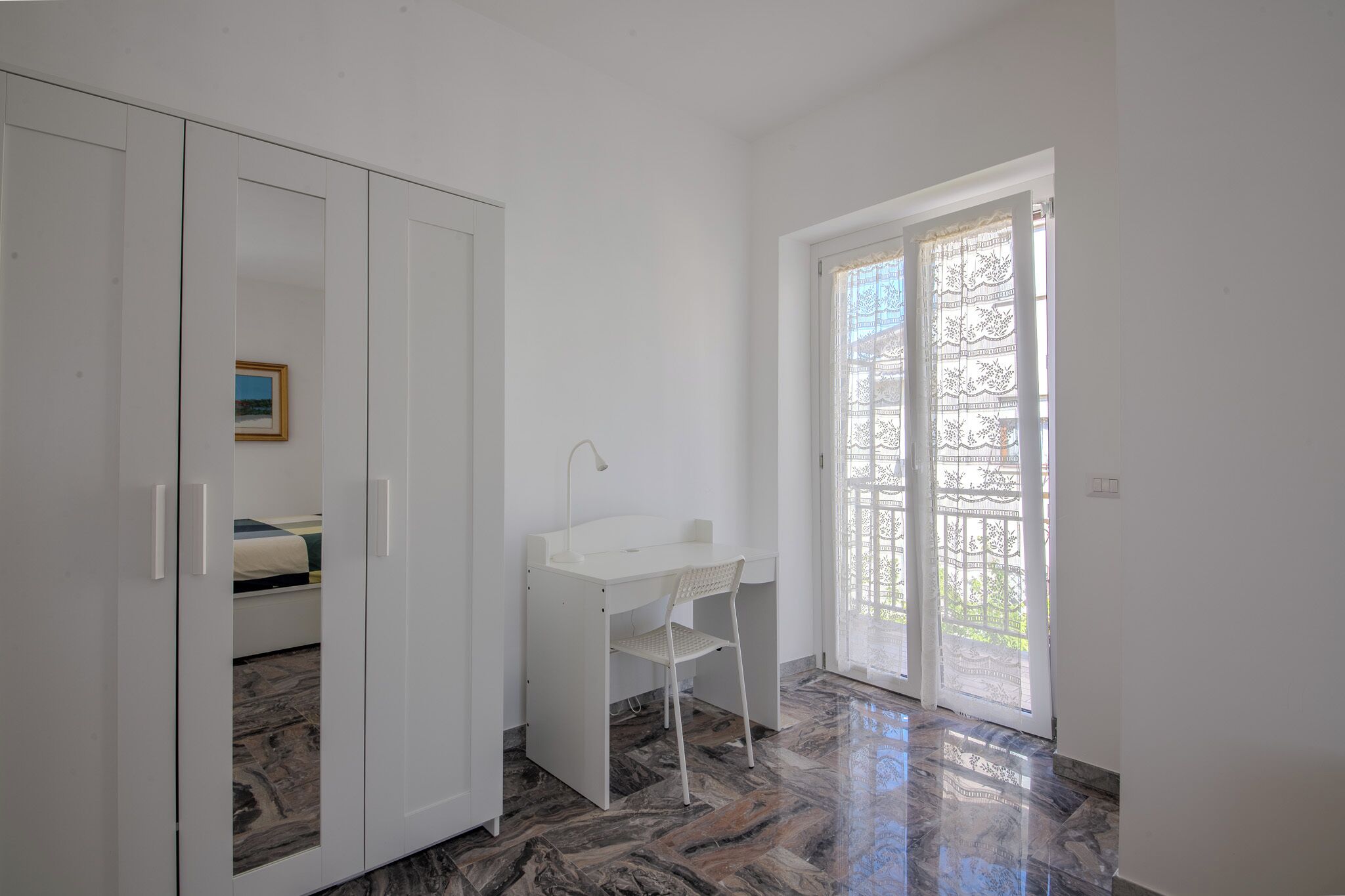 A room with a white desk, a chair, a wardrobe, and a large window with a view of greenery.