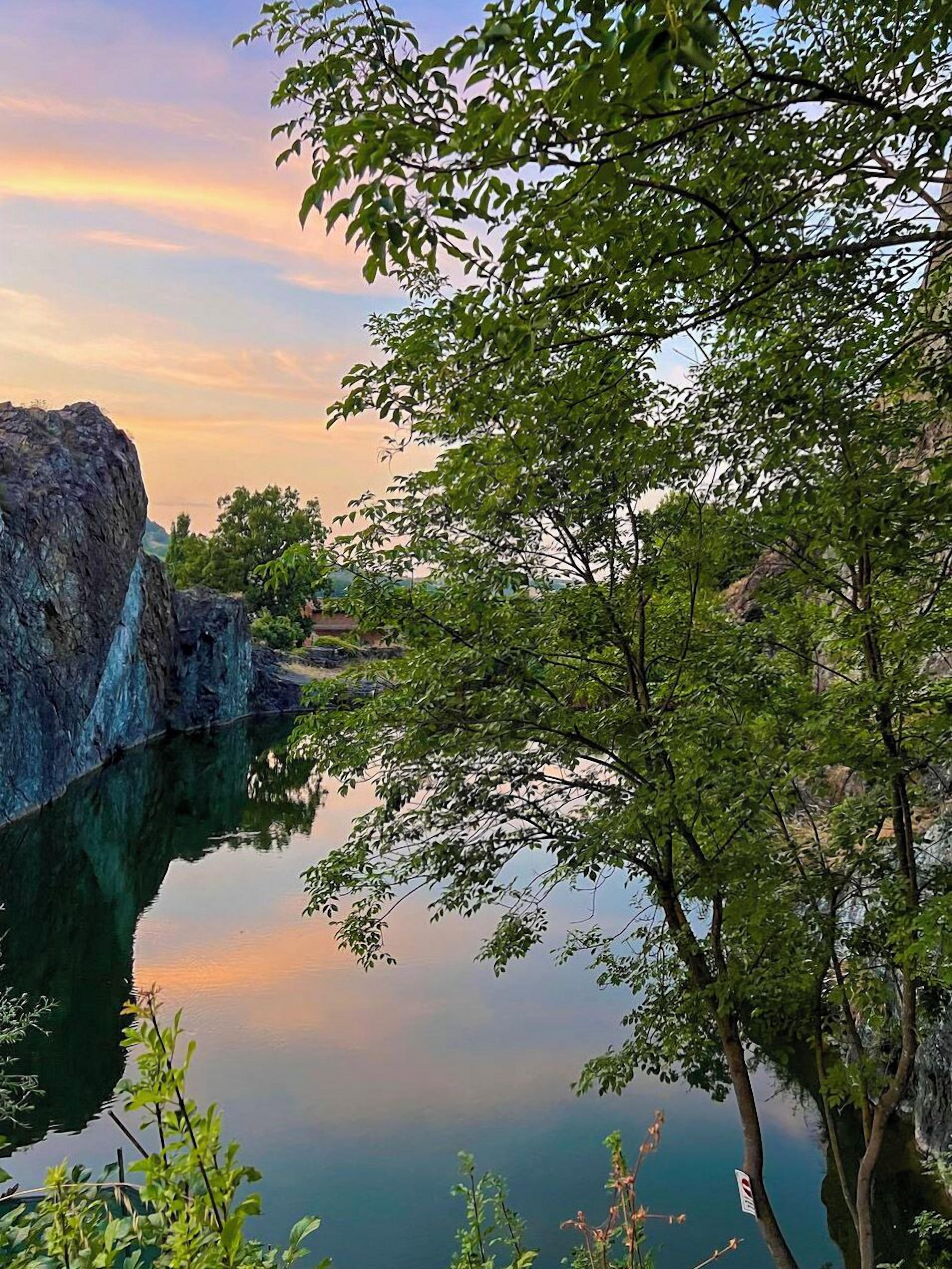A serene landscape with a calm body of water reflecting the sky, surrounded by lush greenery and rocky formations.