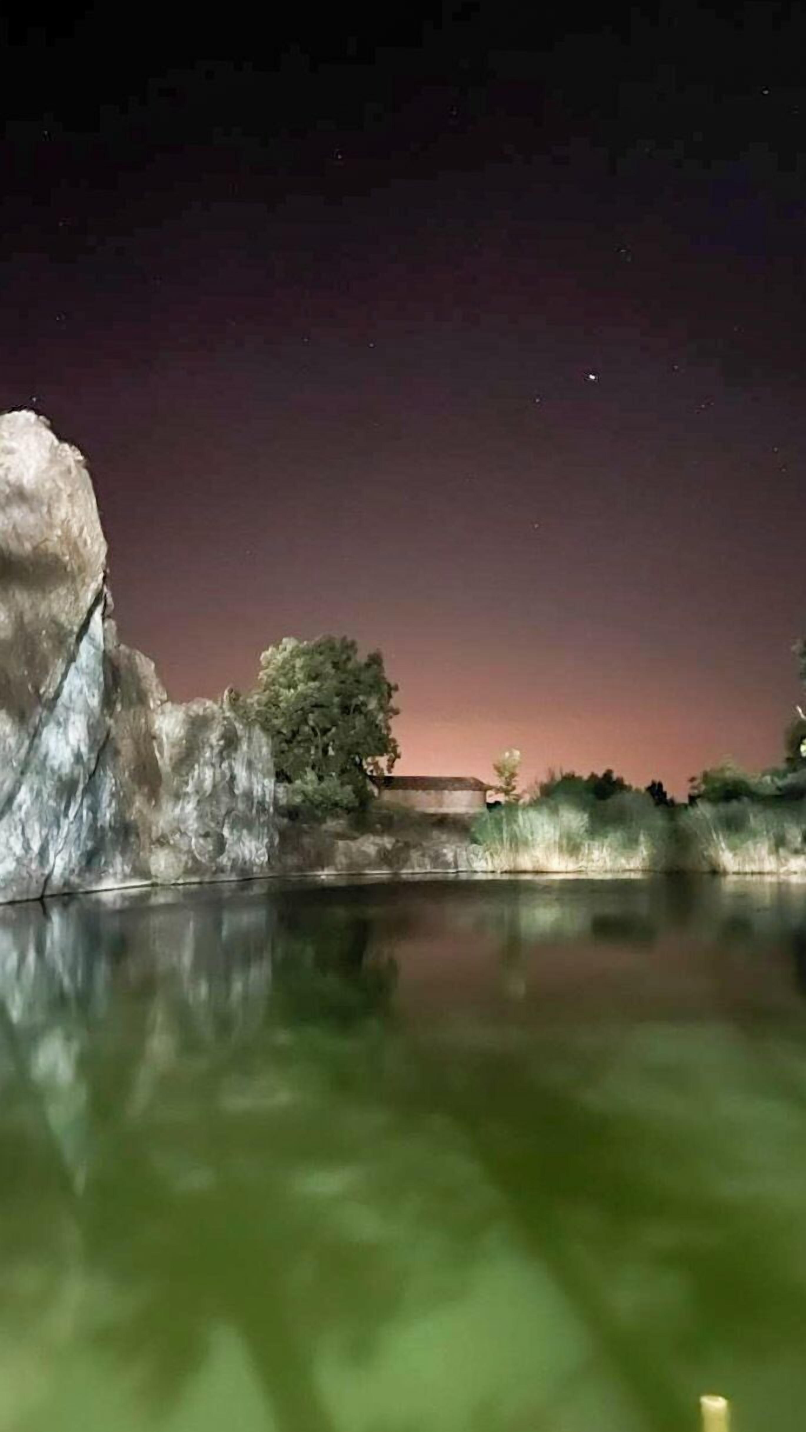 A nighttime view of a calm body of water reflecting a rocky landscape with a tree and a building.