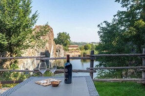 A table with a bottle of wine, two glasses, and a plate of food, set against a backdrop of a natural landscape with a body of water and trees. - Pietra Home (Salsomaggiore Terme)