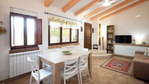 Un coin repas avec une table en bois et des chaises blanches, une cuisine avec un radiateur et un salon avec un canapé et une télévision.