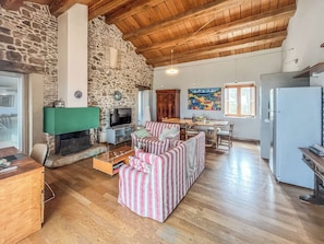 Amplia sala de estar con pared de piedra, techo de madera, chimenea, televisor, mesa de comedor y área de cocina con refrigerador.