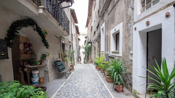 A narrow cobblestone alley with potted plants and a bench, flanked by buildings with balconies and a door. - Il Tasso (Vallerano)