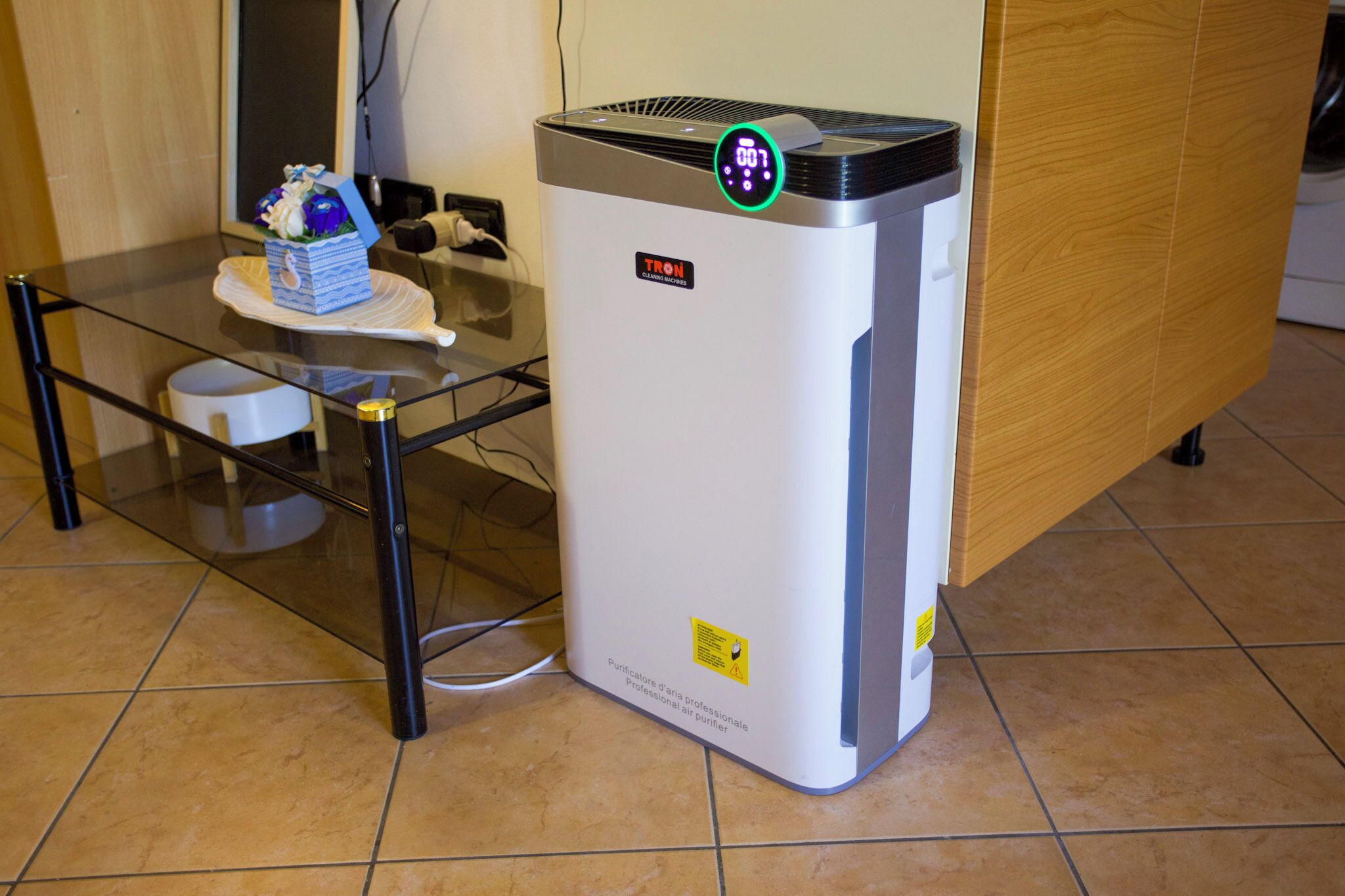 A white air purifier with a digital display on top, placed on a tiled floor next to a glass table with a decorative item.