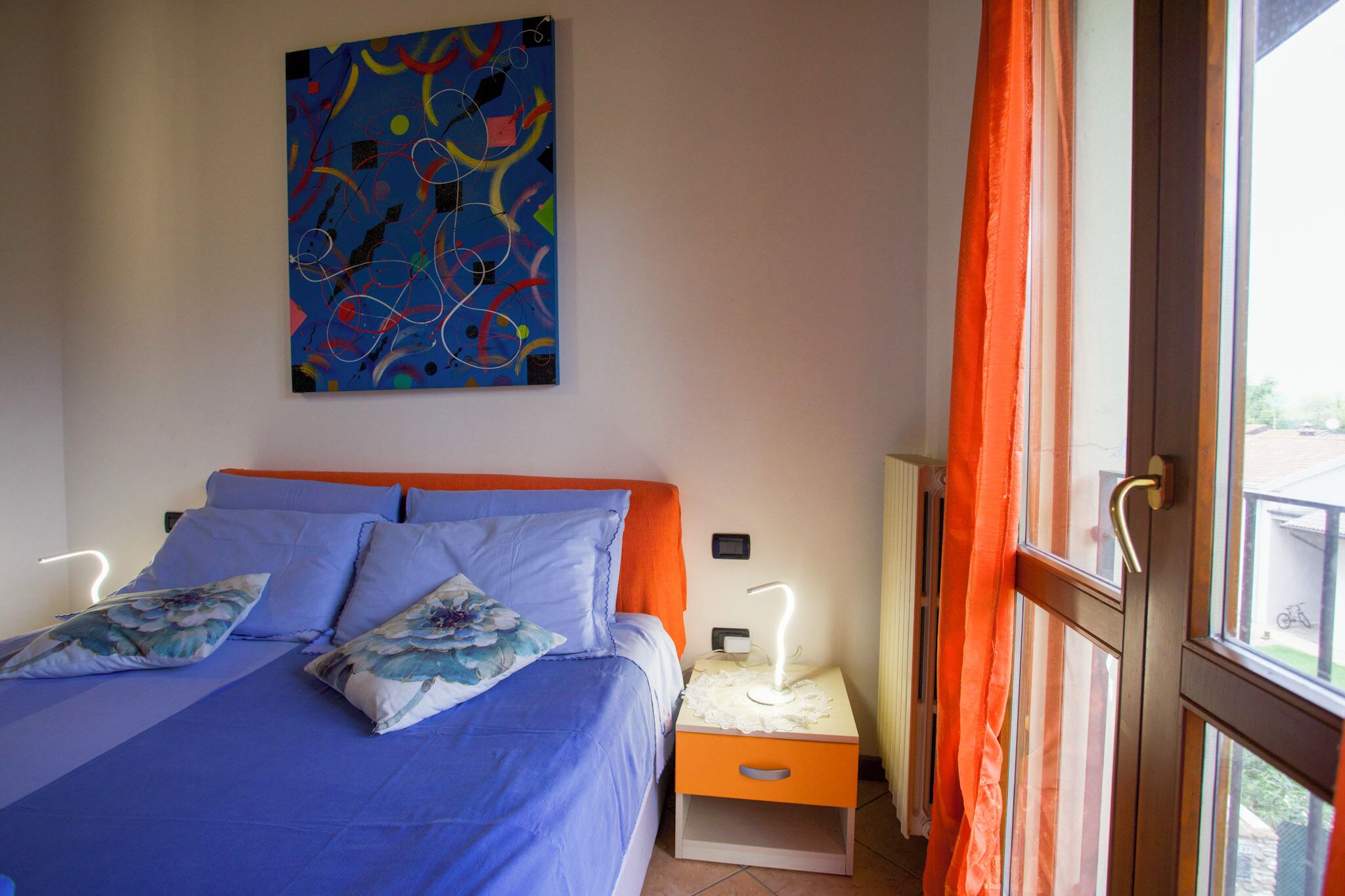 A bedroom with a bed, a nightstand, a lamp, a painting on the wall, and a window with curtains.