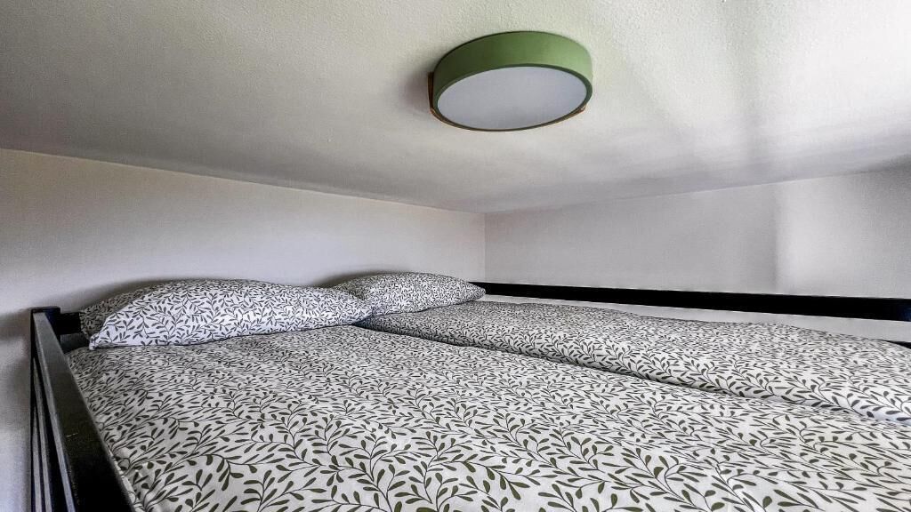 Un lit simple avec une couvre-lit à motifs et des oreillers, un luminaire vert au plafond et des murs blancs.