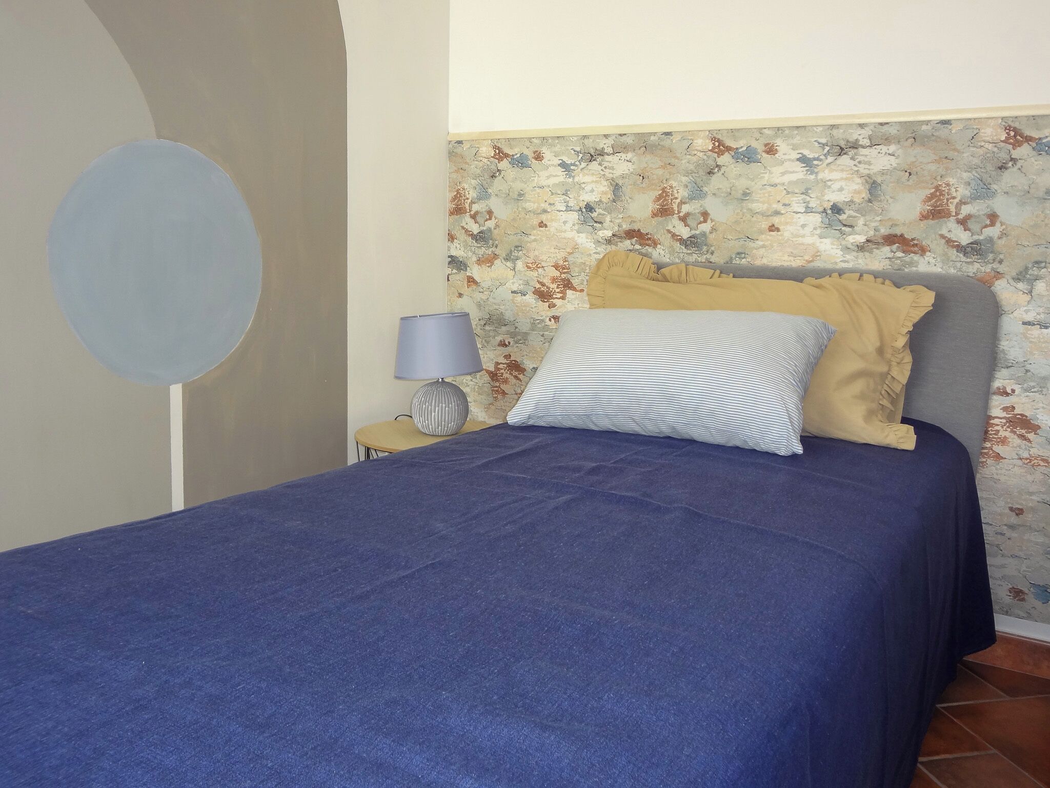 A bed with a blue bedspread, a striped pillow, and a yellow pillow. There is a lamp on a side table next to the bed.