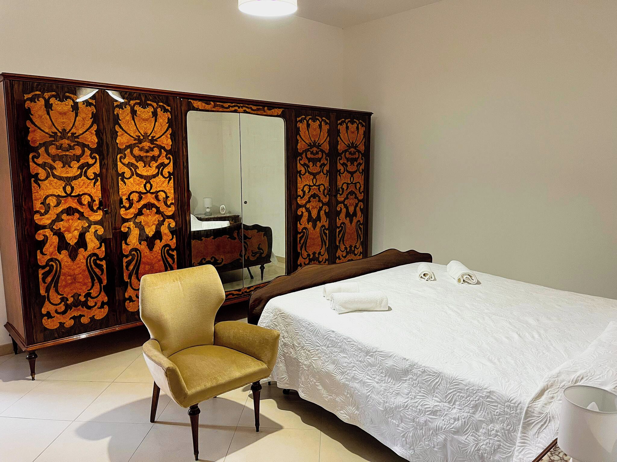 A hotel room with a large bed, a chair, a wardrobe with ornate designs, and a mirror.