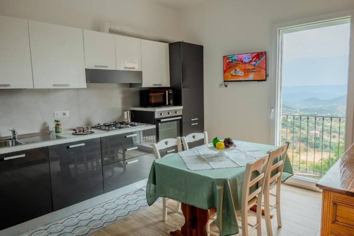 Eine moderne Küche mit schwarzen und weißen Schränken, einer Mikrowelle und einem Herd. Ein Esstisch mit grüner Tischdecke und Stühlen steht daneben. An der Wand ist ein Fernseher angebracht, und ein Balkon mit Blick auf die Landschaft ist zu sehen.