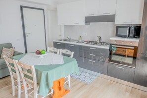 A compact kitchen with modern appliances, a dining area with a table set for two, and a sofa in the adjacent living space. - Sk Barigau (Lanusei)