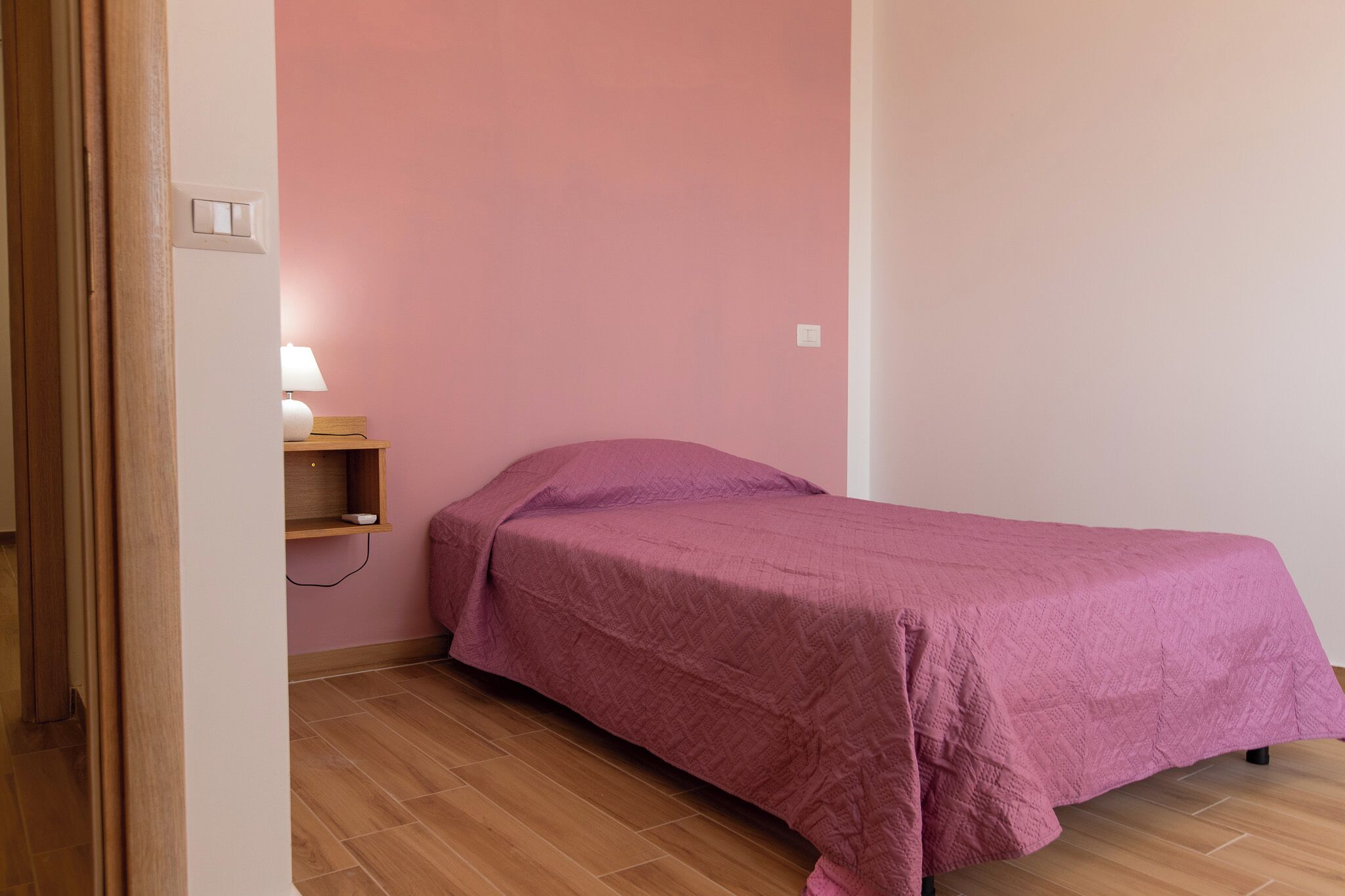 A single bed with a pink bedspread in a room with wooden flooring and a pink wall.
