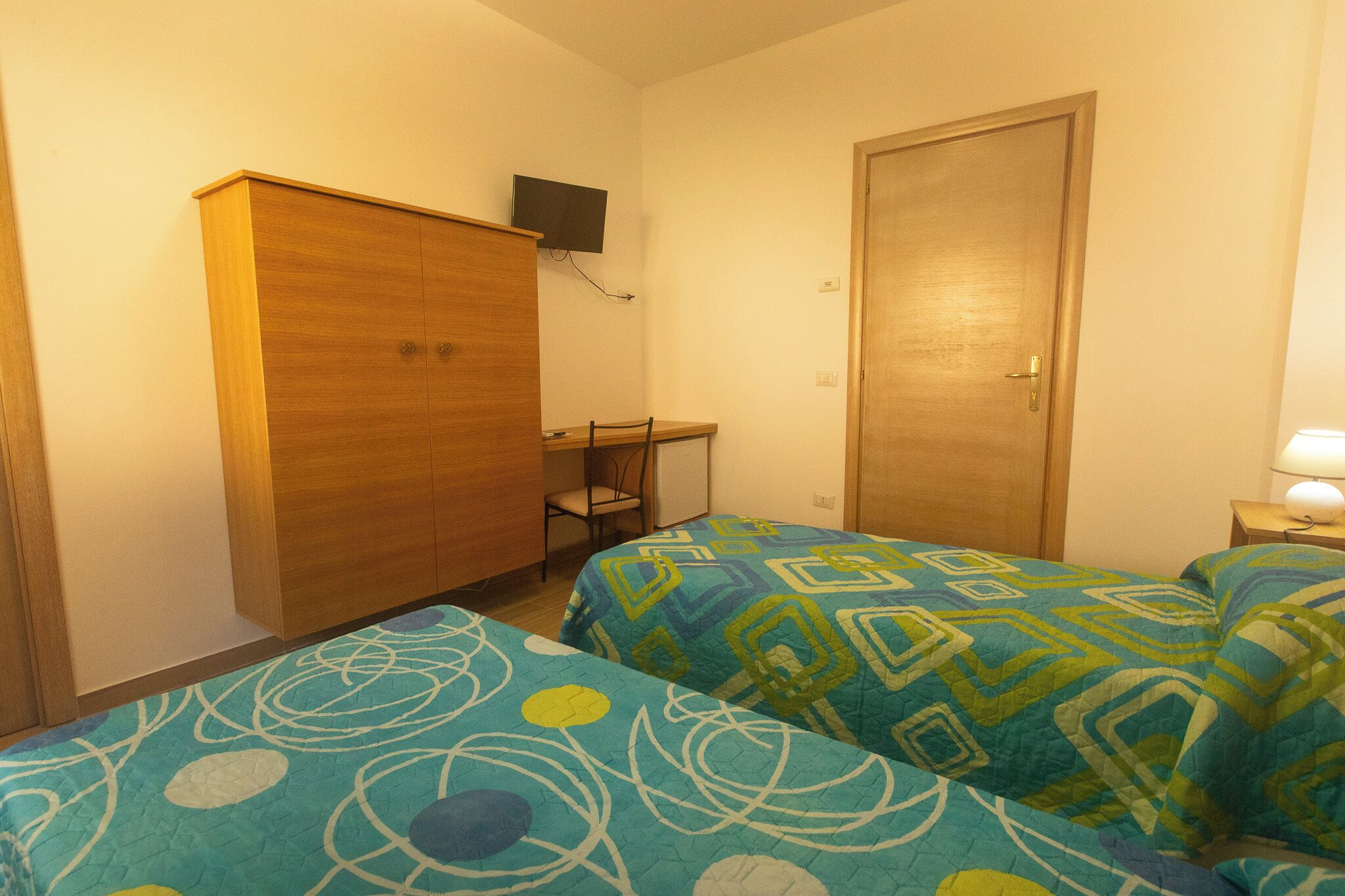 Une chambre avec deux lits, une armoire en bois, un bureau, une chaise et un téléviseur fixé au mur.