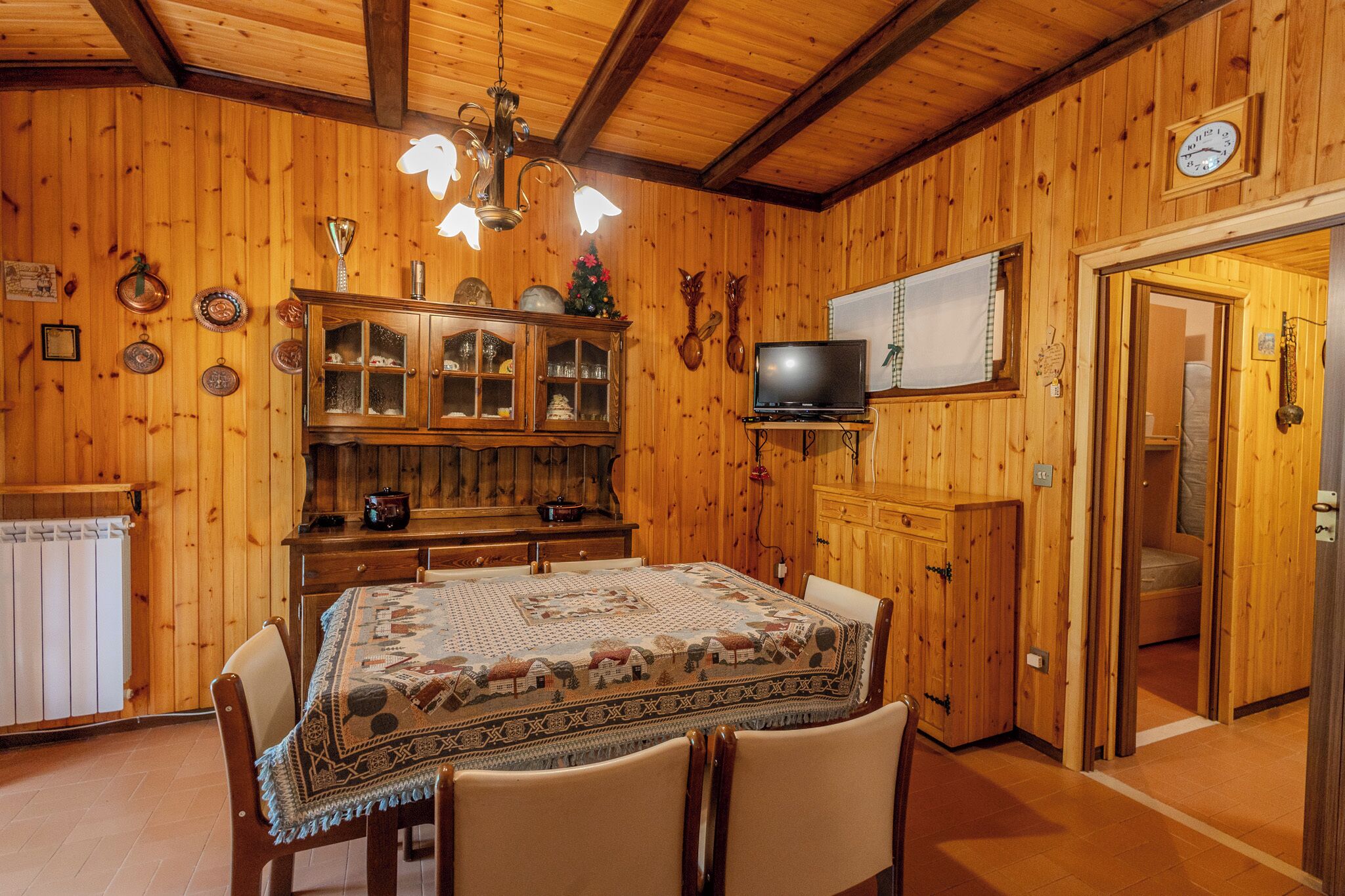 A wooden interior with a dining table, chairs, a chandelier, a television, and a cabinet.
