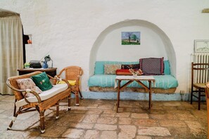 Um quarto aconchegante com piso de pedra, um sofá de vime com almofadas, uma mesa pequena e um sofá azul com manta estampada.