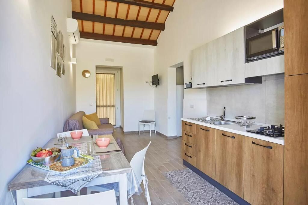 A modern kitchen with wooden cabinets, a microwave, and a sink. There is a dining area with a table set for two, chairs, and a coffee table with fruits and a coffee pot.