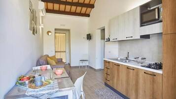 A modern kitchen with wooden cabinets, a microwave, and a sink. There is a dining area with a table set for two, chairs, and a coffee table with fruits and a coffee pot.