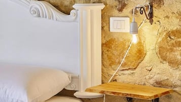 A bedroom with a white bed, a wooden nightstand, a wall-mounted light fixture, and a stone wall.