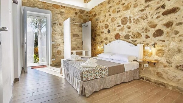 A bedroom with a stone wall, a bed with white linens, a bedside table with a lamp, and a door leading to a balcony. - Casale Sammartano By L.A.Apartments (Ummari)