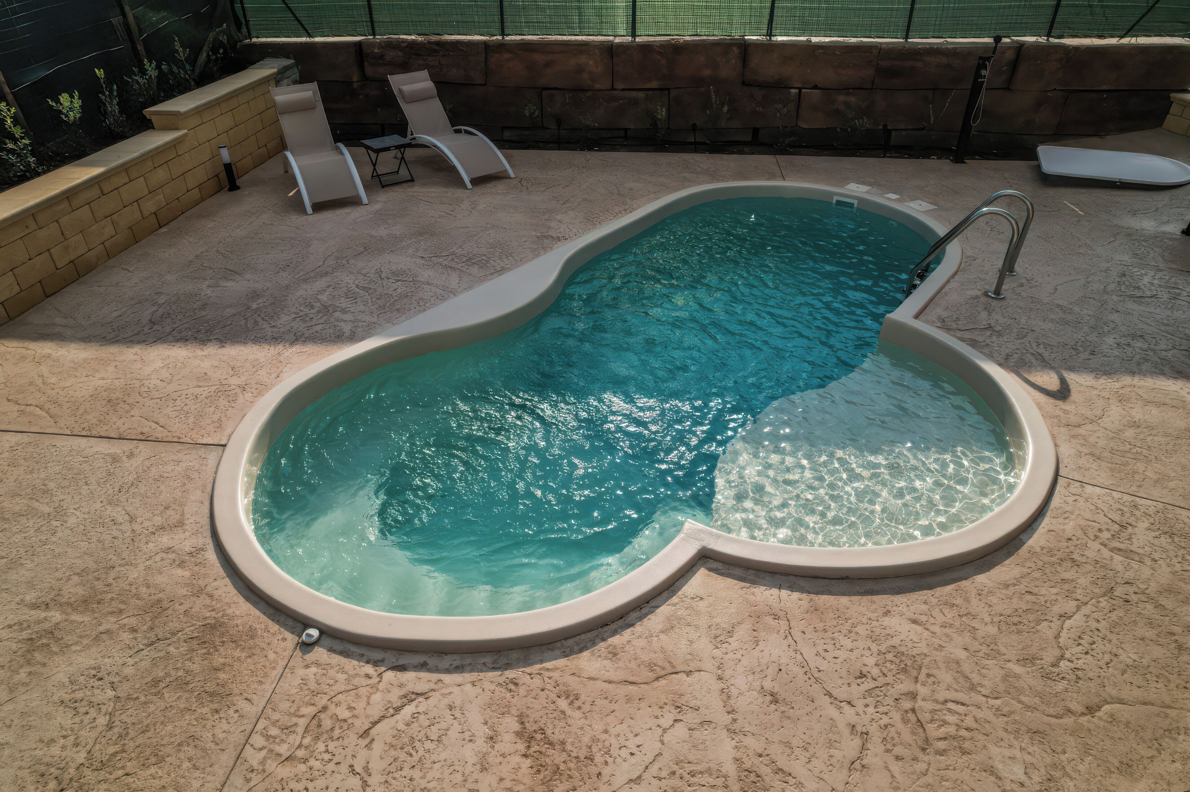 A swimming pool with a unique shape, a stone wall, and a few lounge chairs.