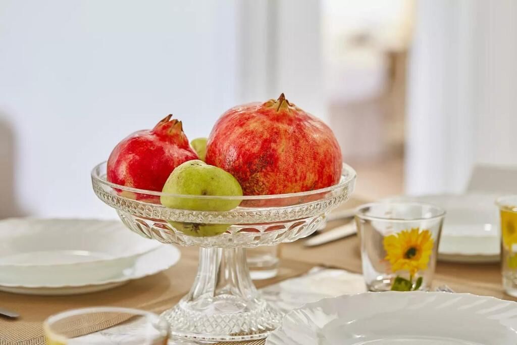 Un cuenco de vidrio transparente sobre una mesa, con una granada, una manzana verde y una fruta amarilla, rodeado de platos blancos y un vaso con diseño de girasol.