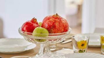 Un cuenco de vidrio transparente sobre una mesa, con una granada, una manzana verde y una fruta amarilla, rodeado de platos blancos y un vaso con diseño de girasol.