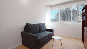 Ein modernes Wohnzimmer mit einem grauen Sofa, einem kleinen weißen Tisch und einem Fenster mit Blick auf Gebäude und Bäume.